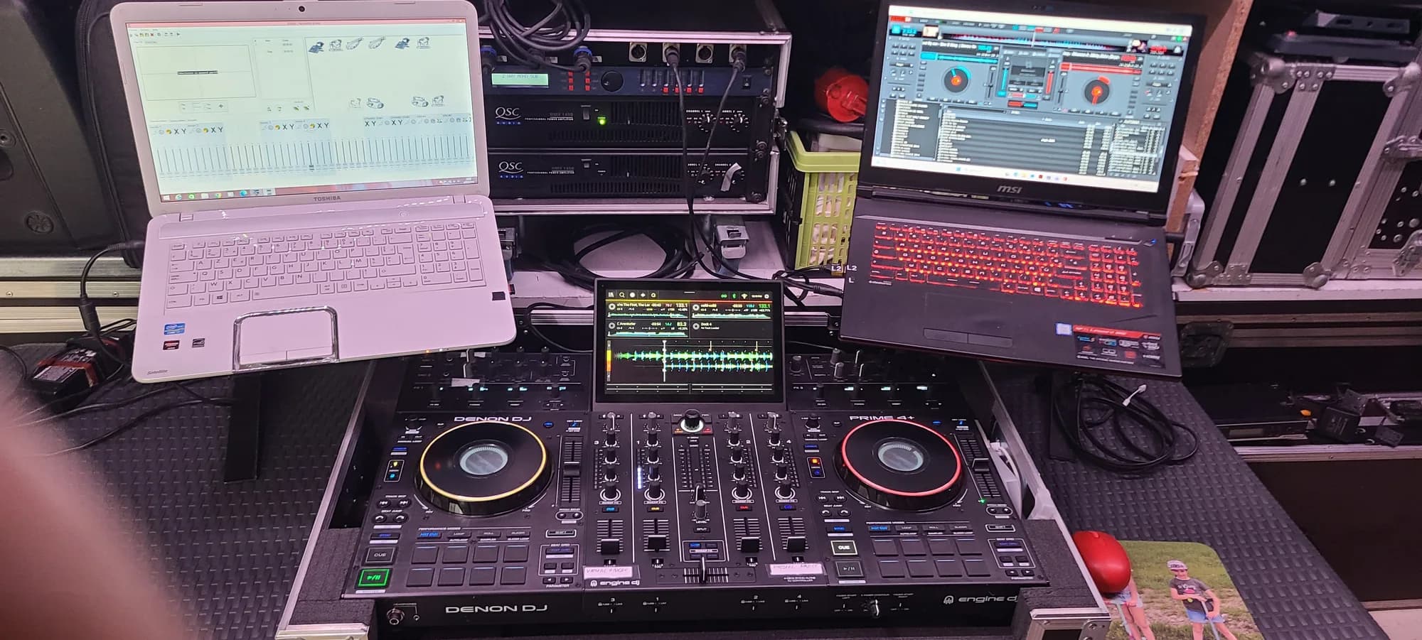Console DJ Denon Prime 4 posée sur un flight case entre deux ordinateurs portables, avec des amplificateurs QSC en rack