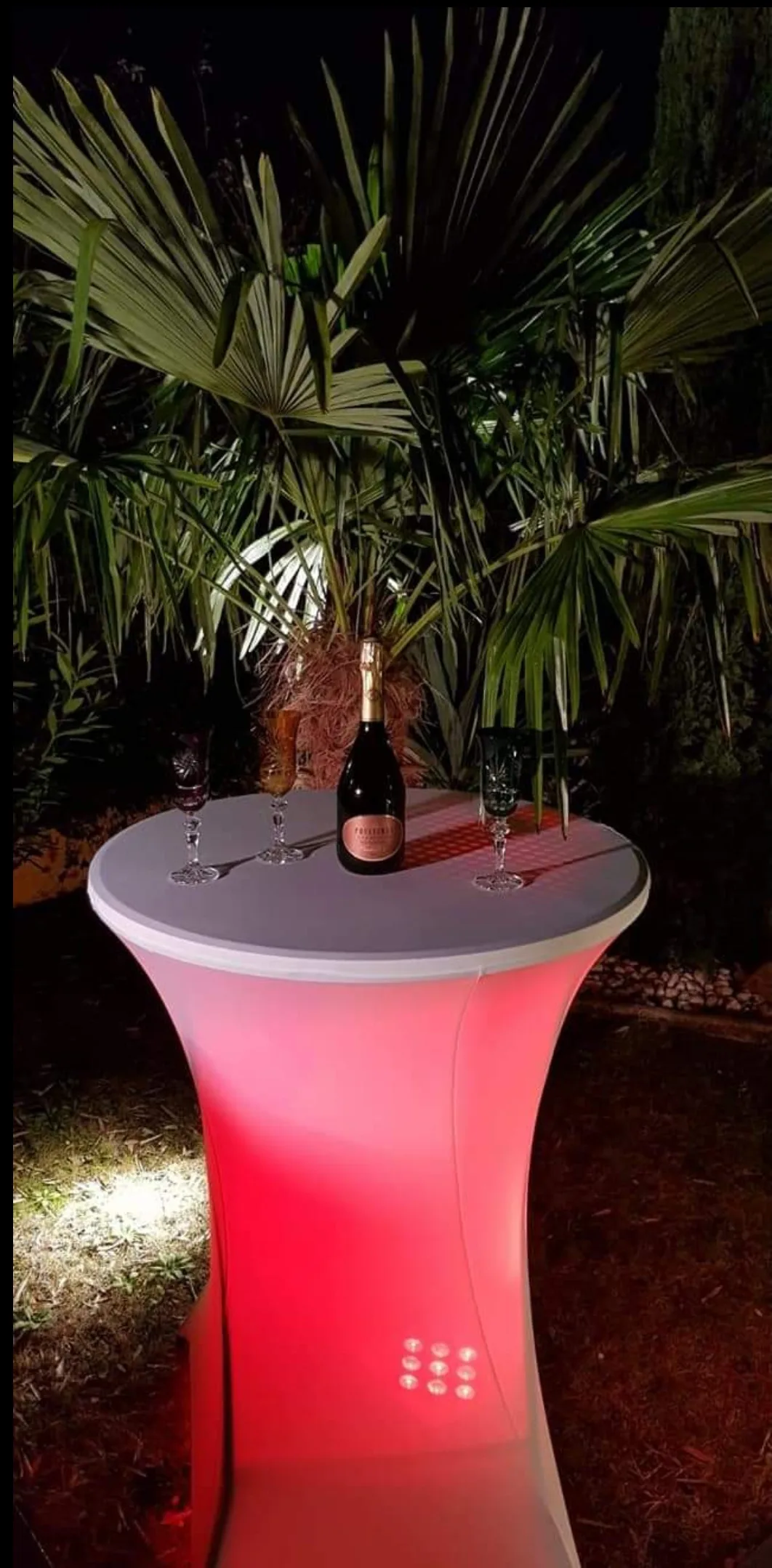 Mange-debout lumineux éclairé en rouge, avec une bouteille de champagne et trois flûtes, devant des palmiers