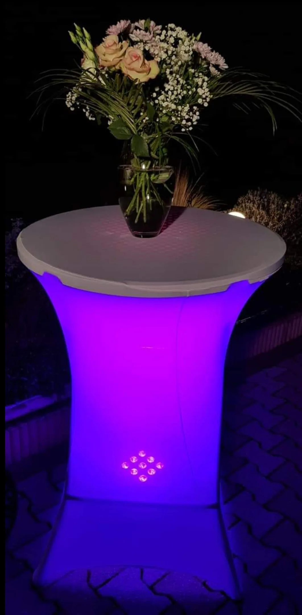 Mange-debout lumineux éclairé en violet, surmonté d'un bouquet de roses et de gypsophile, la nuit