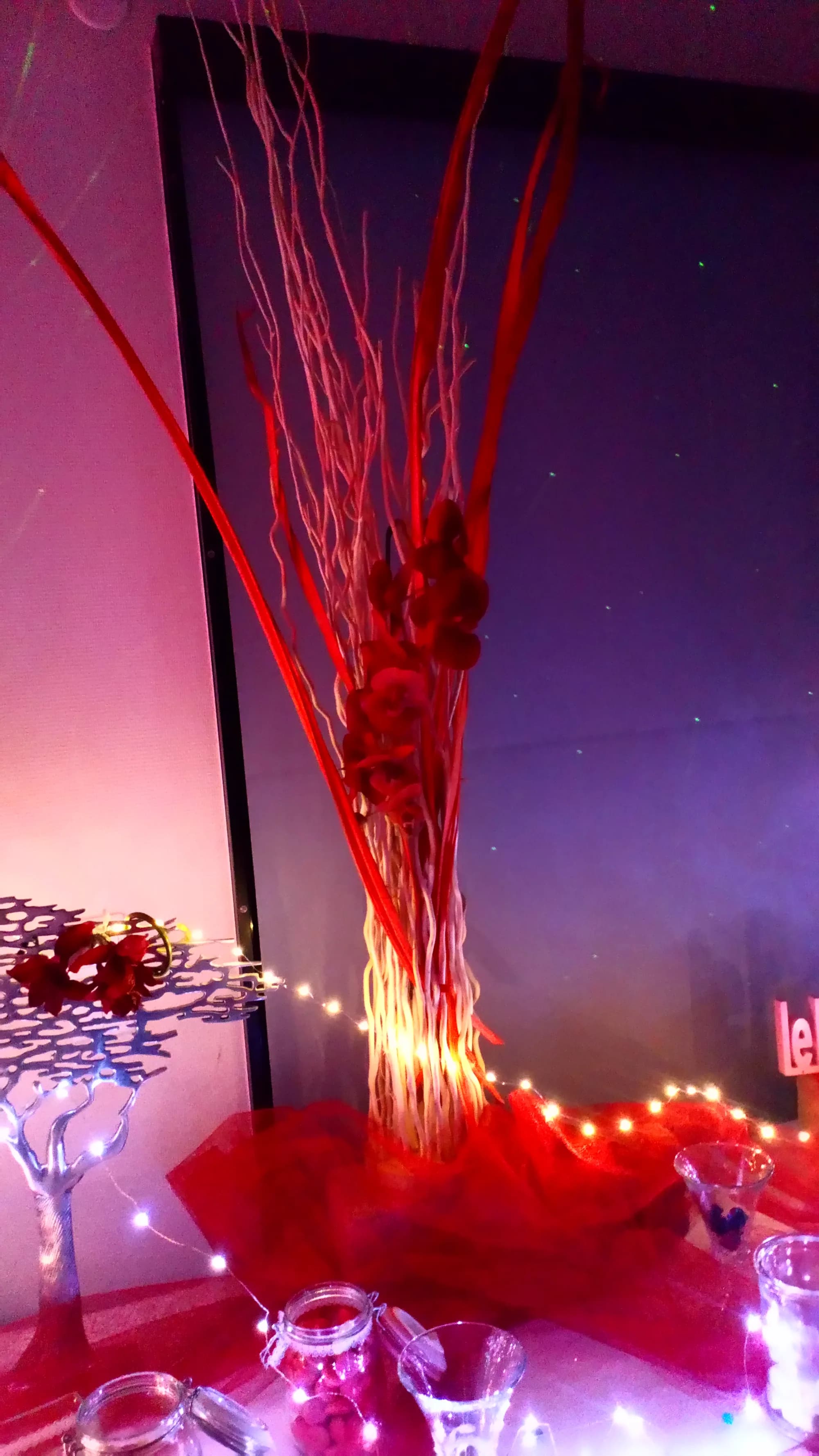 Décoration de table en rouge, branches laquées et orchidées piquées dans une guirlande lumineuse, devant un rideau étoilé