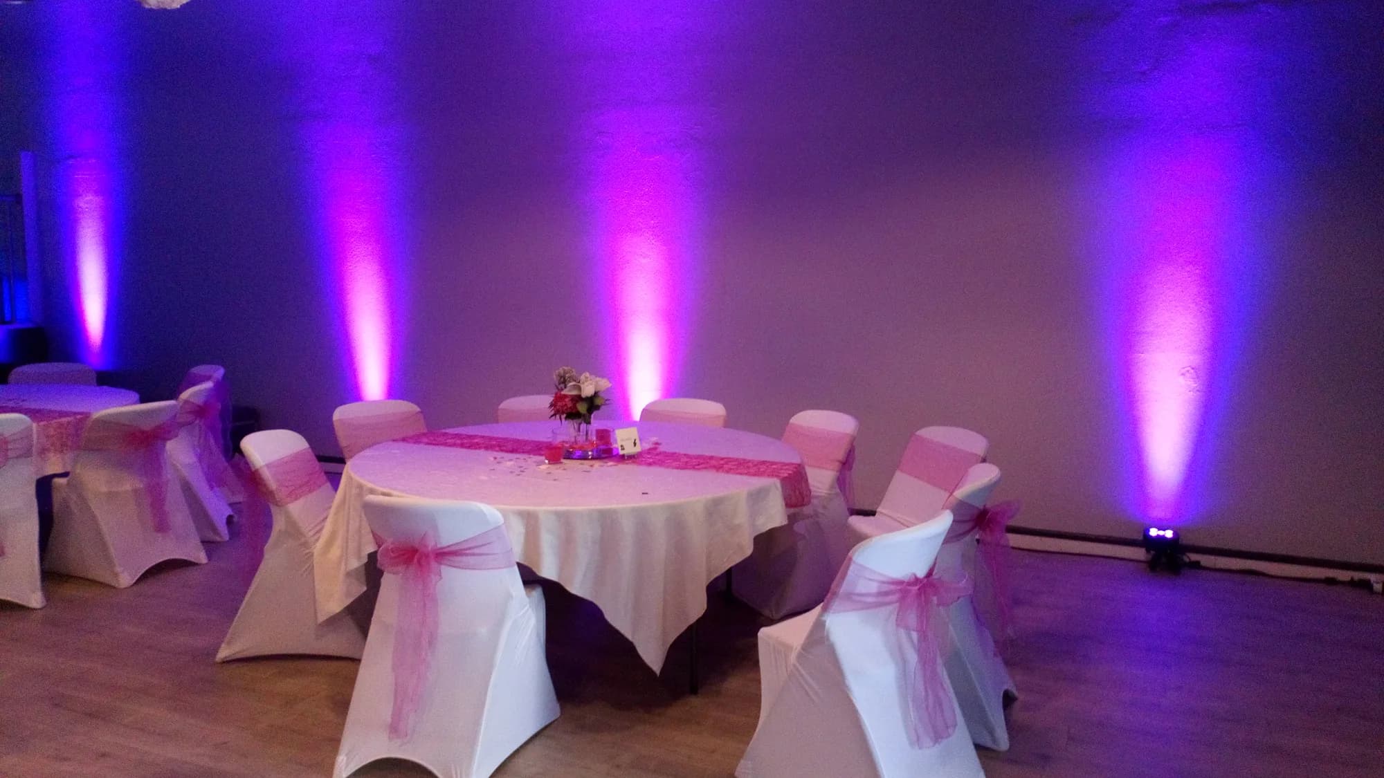 Table ronde dressée avec housses de chaises à nœuds roses, devant un mur éclairé en rose et violet