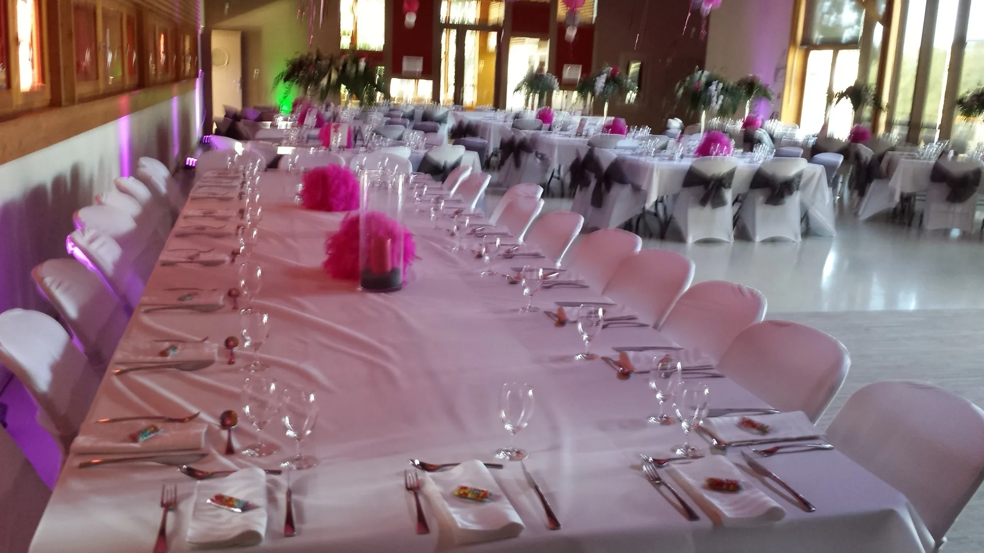Longue tablée de mariage nappée de blanc, housses de chaises à nœuds gris et pompons fuchsia, sous un éclairage violet