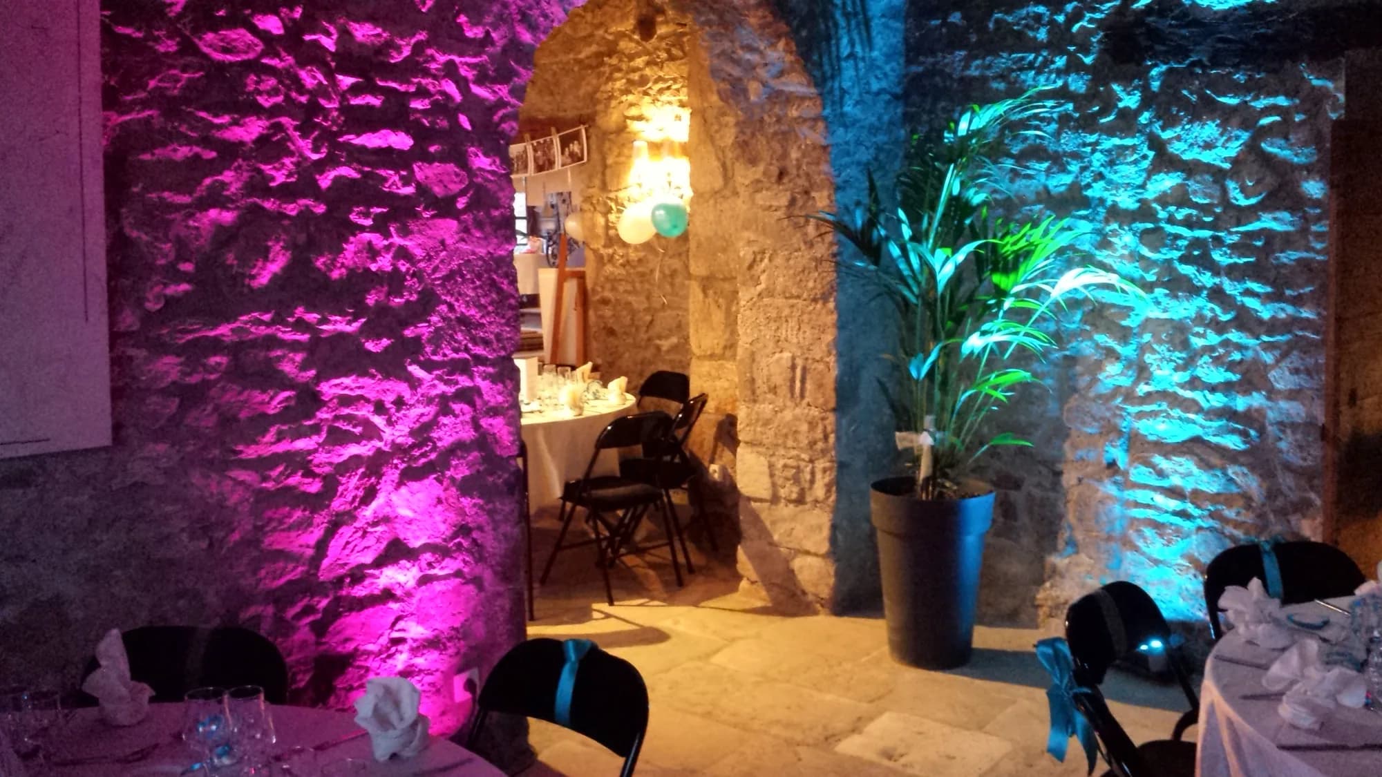 Salle voûtée en pierre dont les murs sont éclairés en magenta et bleu turquoise, tables dressées au premier plan