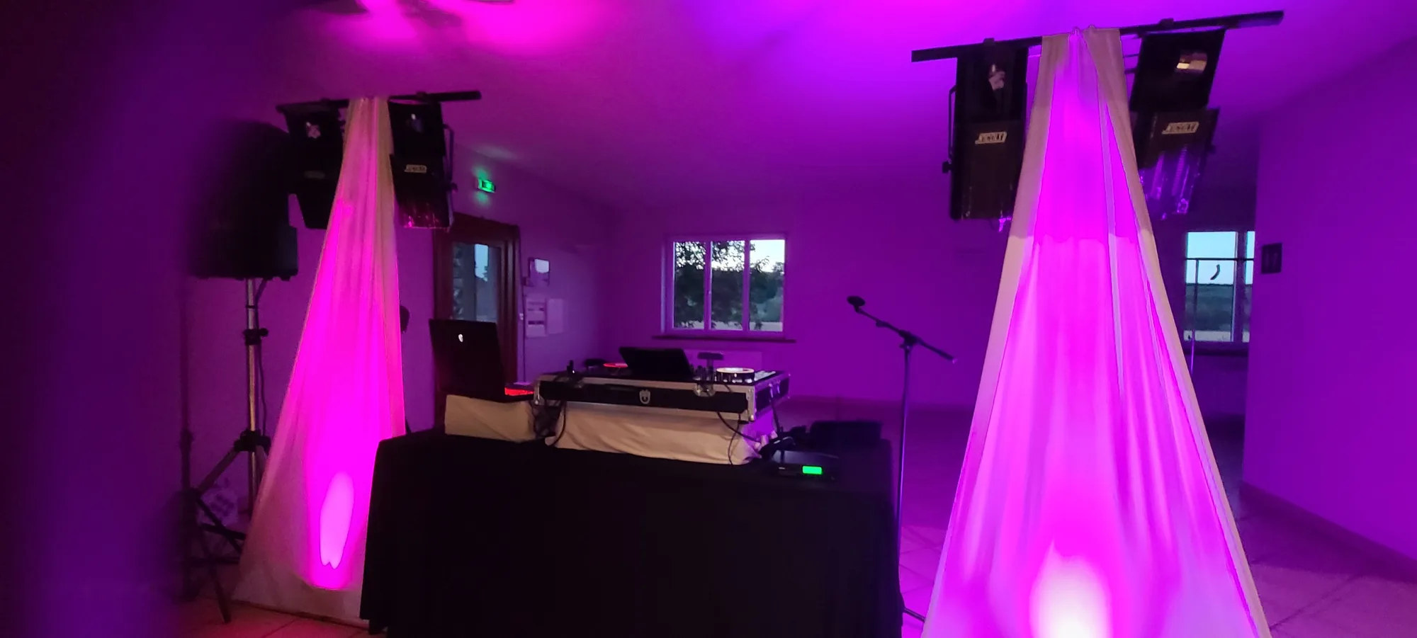 Installation DJ dans une petite salle : table nappée de noir, enceintes sur pieds et deux drapés blancs éclairés en rose